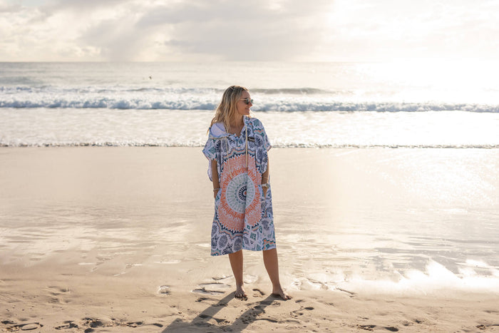 Blue and coral mandala-print hooded towel worn over shoulder at beach, showing sand-free recycled fabric design.