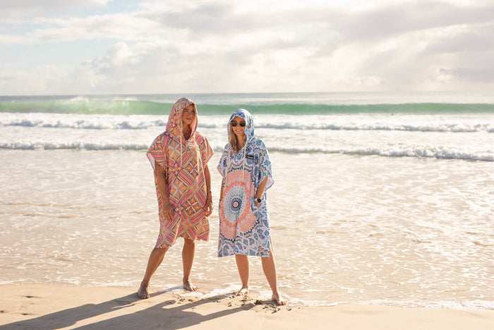 Coral geometric hooded towel worn on beach, paired with blue patterned hooded towel, both featuring drawstring hoods.