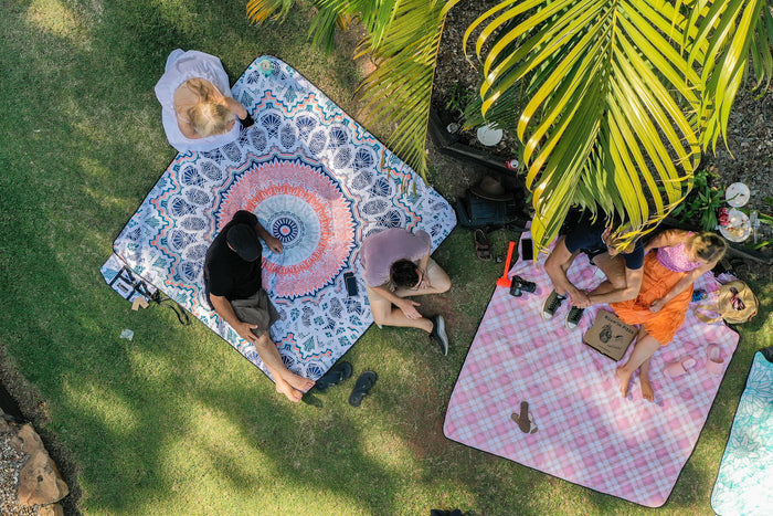 Recycled picnic blanket with blue geometric and coral mandala pattern, displayed outdoors with people lounging on grass.