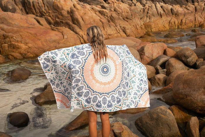 Recycled sand-free beach towel with blue and orange geometric mandala pattern, worn by person at rocky beach