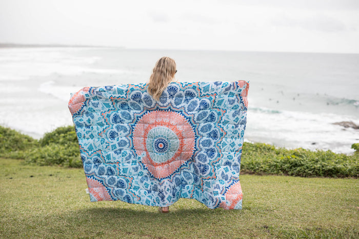 Blue and coral mandala-print sustainable down puffy blanket held up outdoors overlooking ocean.