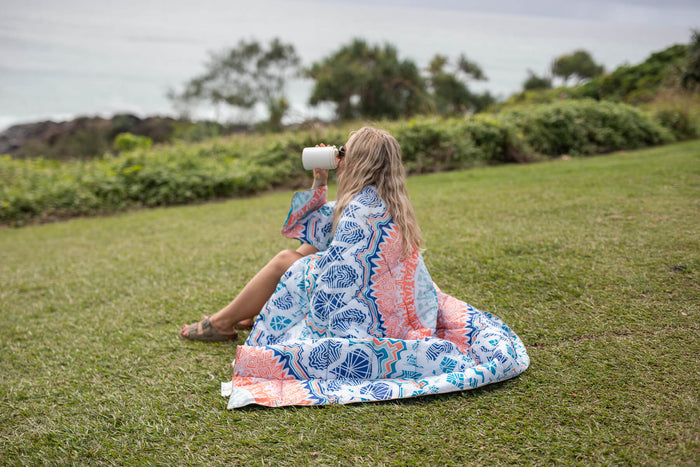 Sustainable down puffy blanket in blue, coral and white geometric print, shown draped over person sitting on grass