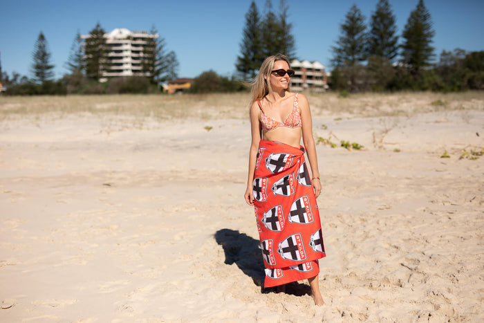 Red AFL beach towel with white cross and shield pattern, draped around woman's waist on sandy beach