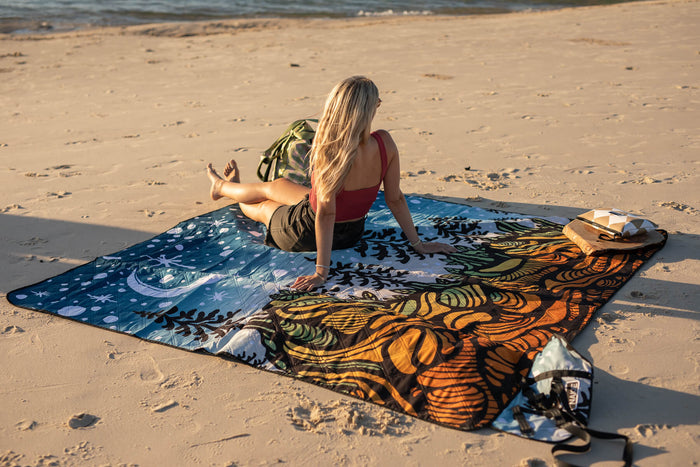 Blue and orange patterned recycled picnic blanket laid on beach sand with woman sitting on it holding a kite.