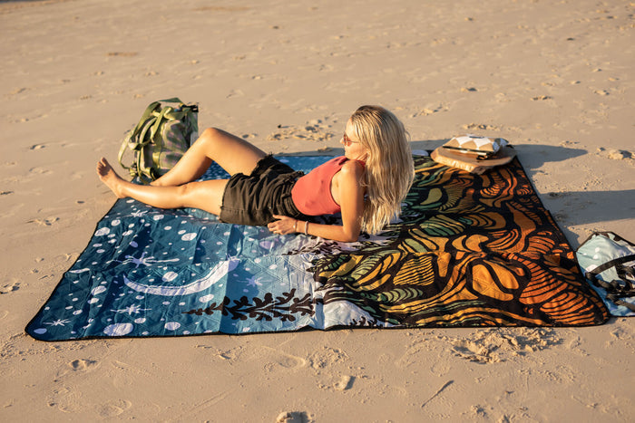 Recycled picnic blanket with blue starry night and orange patterned sections laid on sand beach.