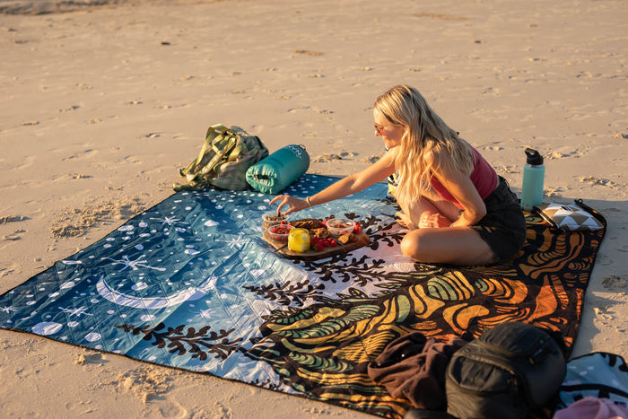 Blue starry-patterned recycled picnic blanket spread on beach sand with woman seated on it.