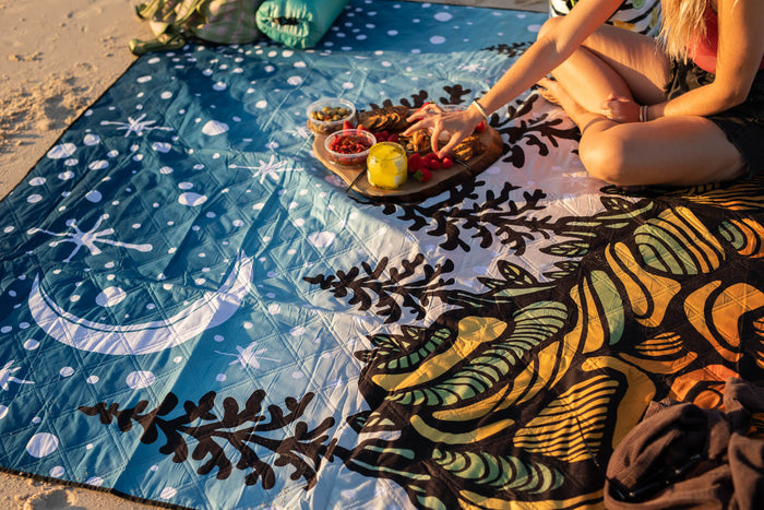 Blue recycled picnic blanket with white starry pattern and botanical motifs, spread on sand with food.