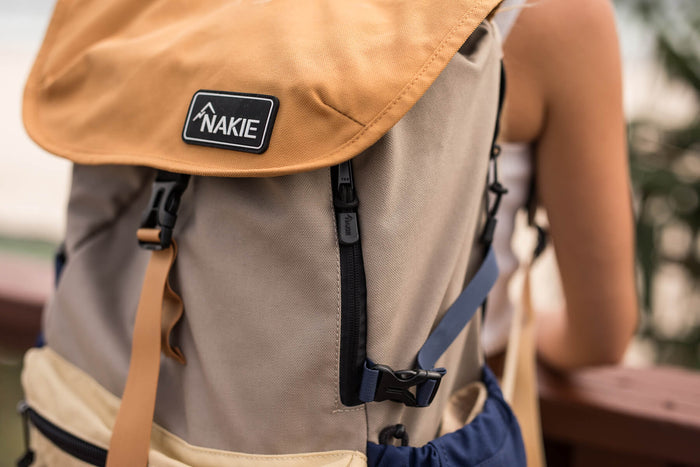 Tan and beige color-block recycled travel backpack with black straps and NAKIE logo patch, front view.