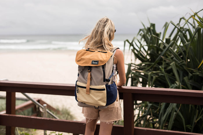 Tan, gray, and navy colorblock recycled backpack with branded label, worn by woman overlooking beach.