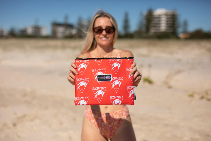 Red Sydney Swans AFL beach towel with repeating swan logo and text pattern, held up on sandy beach