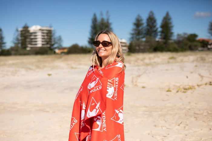 Red Sydney Swans beach towel with white logo print, draped over woman's shoulders on sandy beach