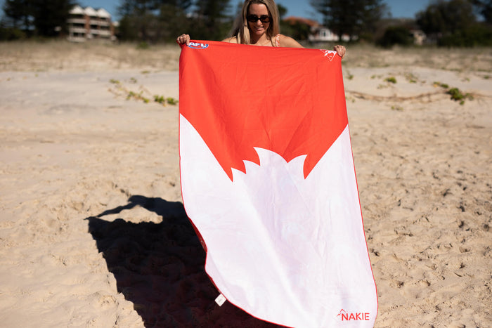 Red and white Sydney Swans AFL beach towel with swan logo, held on sandy beach