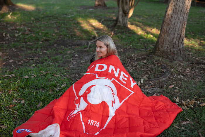 Red and white Sydney Swans AFL puffy blanket with logo and "1874" text, draped over person outdoors