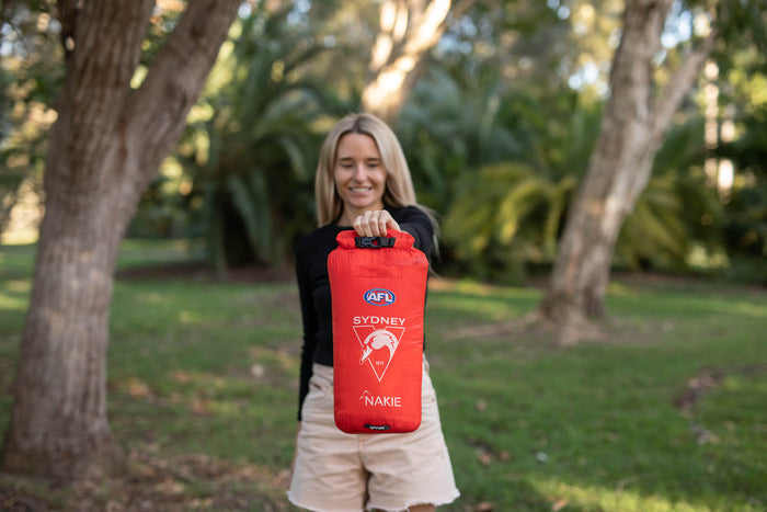 Red AFL Sydney Swans waterproof dry bag with roll-top closure and Nakie branding, held by person outdoors.