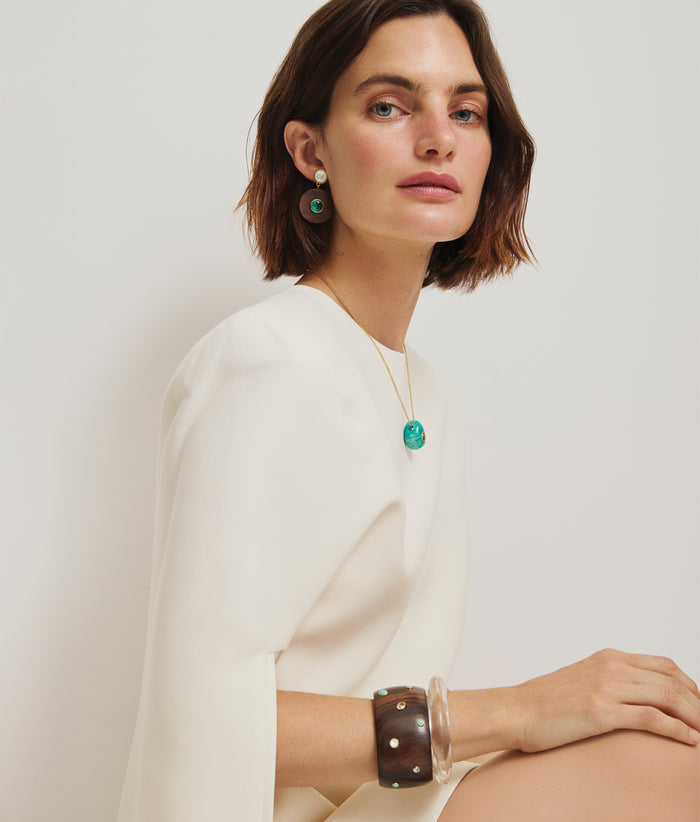 Model in white cape-dress wears Taj Disc Earrings in Acacia Wood, Constance Necklace and Lana Cuff