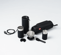 Portable travel coffee kit: grinder, metal filter, scoop, small cup, power cord, and zippered black case on white background