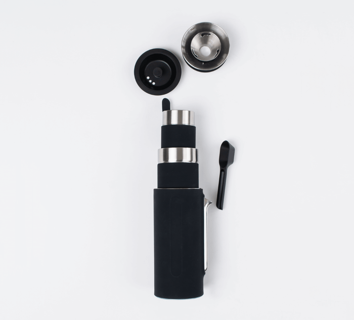 Black Pakt Coffee Kit with a cylindrical main body, a detachable lid, a metal filter, and a small black scoop, all arranged on a white background.