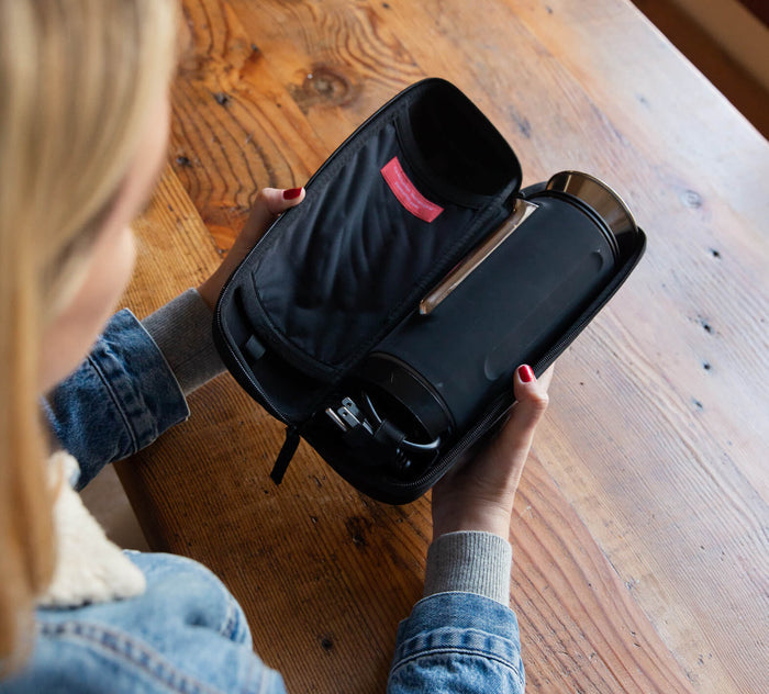 Person holding open black zip carry case with a stainless insulated travel mug inside, over a wood table.