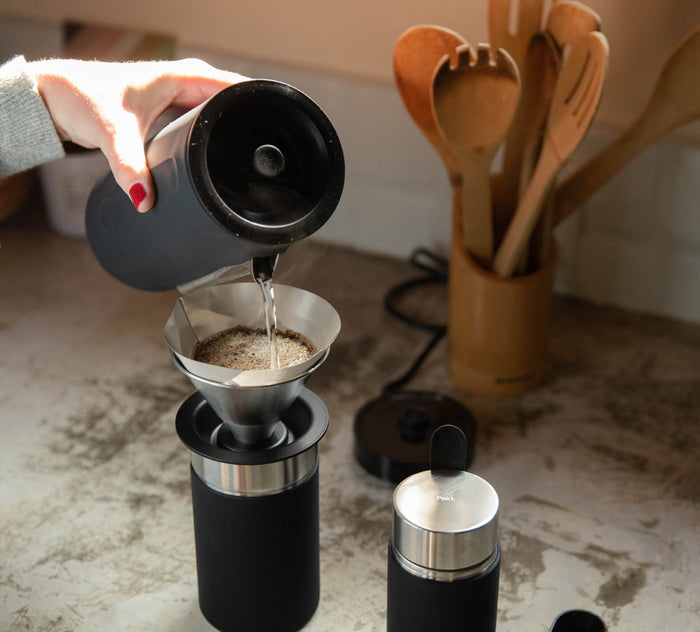Pouring hot water from a black kettle into a coffee dripper over a black Pakt Coffee Kit container on a kitchen counter with wooden utensils in the background