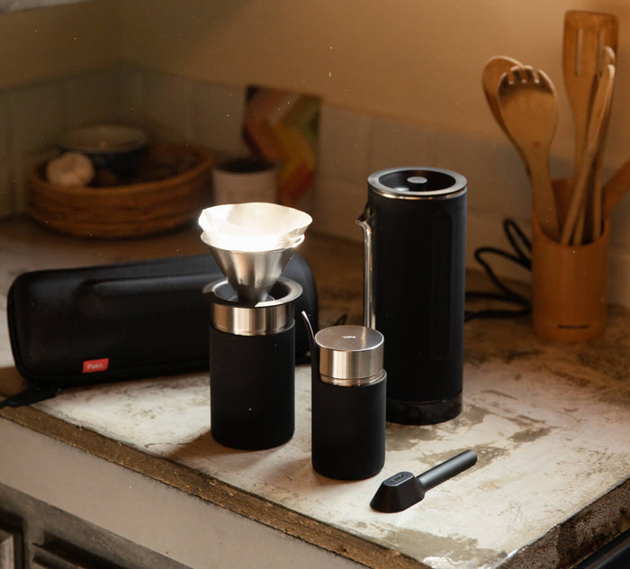 Manual coffee grinder, metal pour-over dripper and travel mug on a kitchen countertop with wooden utensils behind.