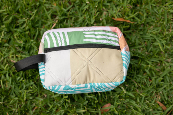 Quilted toiletry bag with multicolored zebra stripe panels in green, turquoise, and beige with black zip closure.