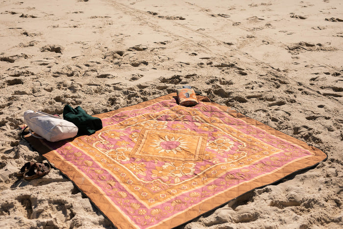 Pink and orange patterned recycled picnic blanket spread on beach sand with person lying down and orange bag nearby