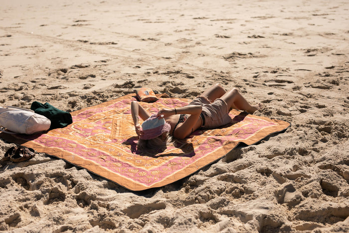 Orange and pink geometric patterned recycled picnic blanket laid on beach sand with person relaxing on it