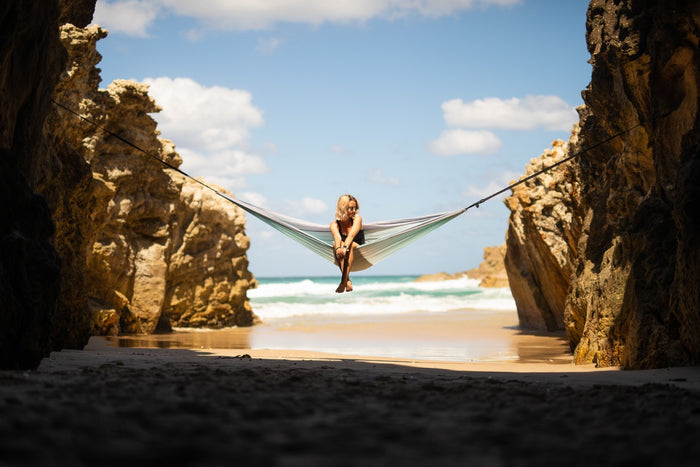 Twilight Blue recycled hammock with straps suspended between coastal rock formations above sandy beach