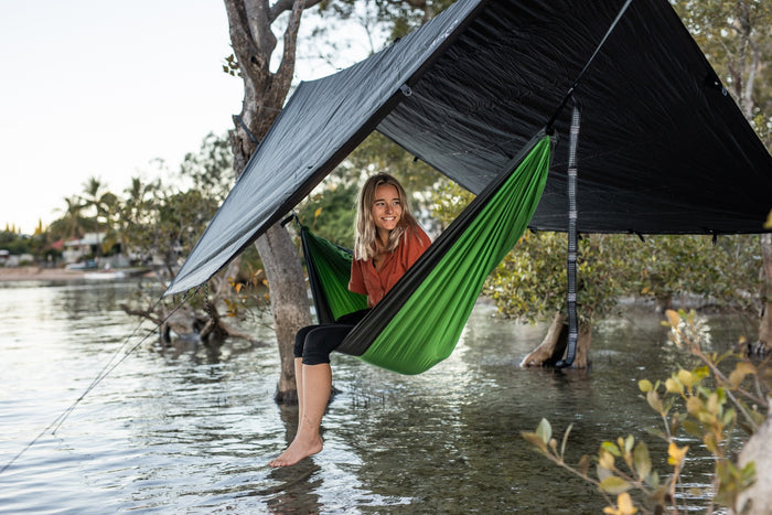 Ultralight tarp in grey with black underside suspended over green hammock near water with trees.