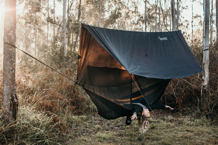 Ultralight tarp in navy blue with burnt orange underside, pitched between trees in forest setting with integrated bug net.