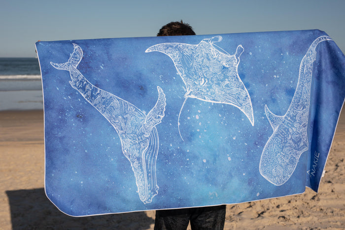 Blue beach towel with white decorative manta ray and stingray illustrations, sand-free texture, held at seaside.