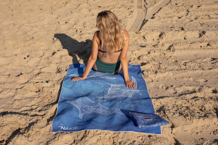 Blue recycled beach towel with white whale and sea creature designs, displayed on sand.