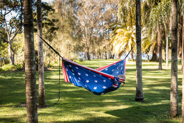 American flag printed recycled hammock with red stripe suspended between two trees in park setting.