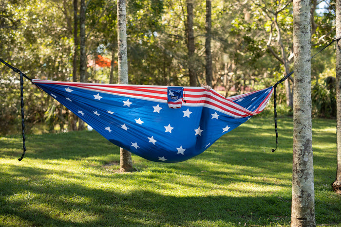 Blue camping hammock with white stars and American flag stripe detail, suspended between two trees with black straps.