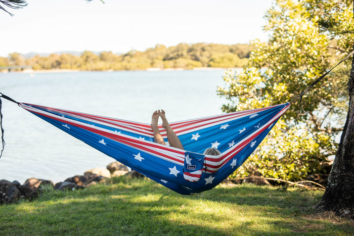 Blue and red American flag-patterned camping hammock with person lounging, strung between trees by lakeside.