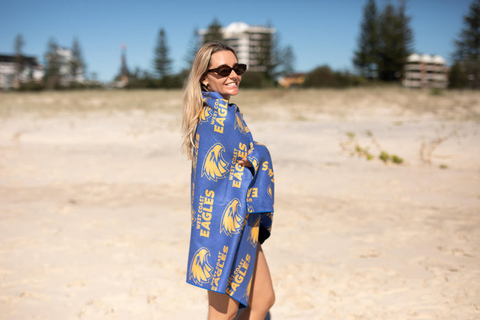 Blue and gold West Coast Eagles AFL beach towel with repeating eagle logo and text, worn over shoulder on sandy beach