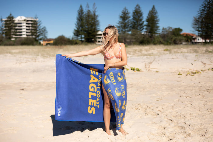 West Coast Eagles blue beach towel with yellow logo text and shell pattern, held open on sand.