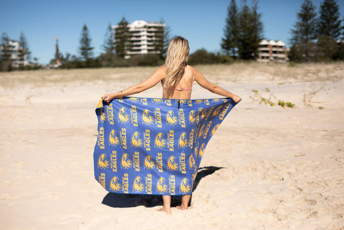 Blue beach towel with West Coast Eagles logo and text pattern, held open by woman on sand.