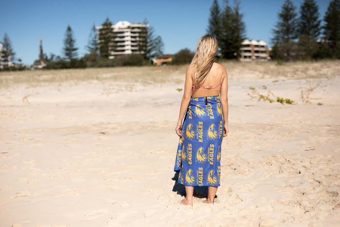 Blue AFL West Coast Eagles beach towel with yellow logo repeat, draped over woman's shoulder at beach