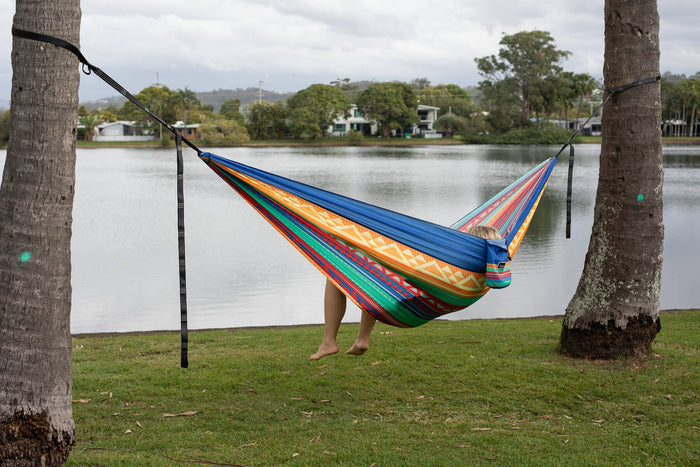 Colorful striped recycled hammock with black straps hung between two trees overlooking water and houses.