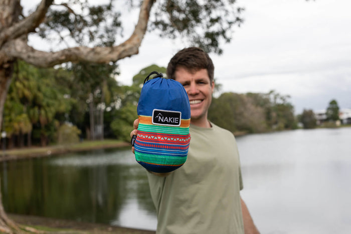 Blue recycled hammock with colorful striped pattern and carrying straps, displayed outdoors by water.