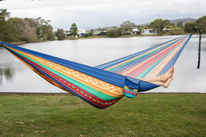 Multicolored striped recycled fabric hammock with blue trim, suspended over grass with water and trees in background