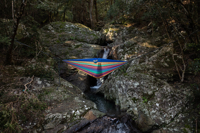 Multicolored striped recycled hammock suspended between rocks over a forest stream with attached straps.
