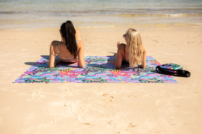 Colorful rainbow floral sand-free beach towel spread on sand with two people sitting on it by the ocean.
