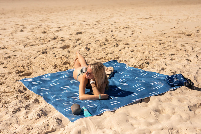 Blue recycled beach blanket with white wave pattern, laid flat on sand with person lounging on it