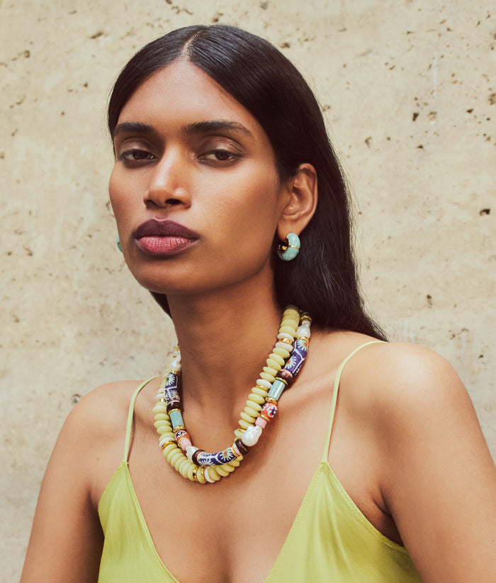 Model on tan stone wall wears yellow top with Zellige Hoops, Souvenir and Laguna Necklaces